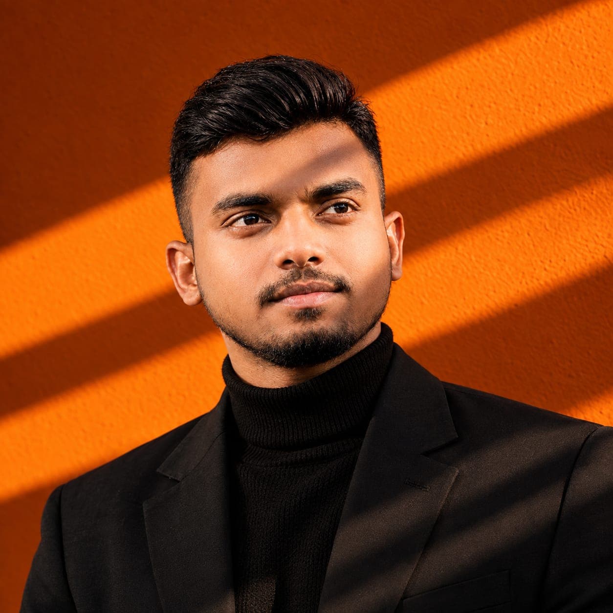 Portrait of Sani in a dark blazer and turtleneck against an orange wall